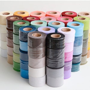 Puede incluir: Surtido de rollos de tela de colores, cada uno envuelto en plástico transparente. Los rollos están apilados en filas, creando una visualización visual de varios colores y texturas.