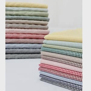 Puede incluir: Una pila de cuadrados de tela doblados con un patrón de cuadros vichy pequeños en varios colores, incluyendo amarillo, verde claro, salvia, beige, rosa, rojo, azul claro y azul marino, dispuestos en dos montones sobre una superficie blanca.