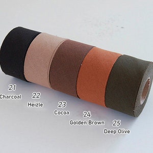 May include: A cylindrical roll of fabric tape featuring five distinct colour segments labelled with numbers and names: 21 Charcoal, 22 Heizle, 23 Cocoa, 24 Golden Brown, and 25 Deep Olive. The material has a matte, textured finish across the entire length.