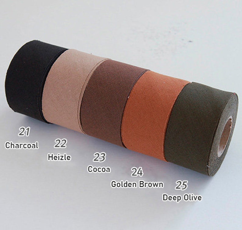 May include: A cylindrical roll of fabric tape featuring five distinct colour segments labelled with numbers and names: 21 Charcoal, 22 Heizle, 23 Cocoa, 24 Golden Brown, and 25 Deep Olive. The material has a matte, textured finish across the entire length.