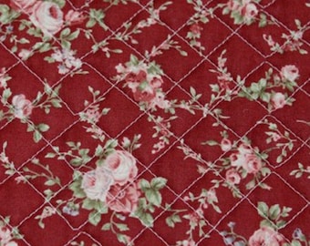 May include: Red quilted fabric with a white floral pattern. The fabric features a repeating pattern of small white flowers and leaves on a red background. The fabric is quilted with a diamond pattern.