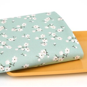 May include: A folded piece of light mint green fabric featuring a repeating pattern of small white cherry blossom flowers on dark brown stems. The fabric is layered on top of a smooth, solid mustard yellow rectangular tray with rounded edges.