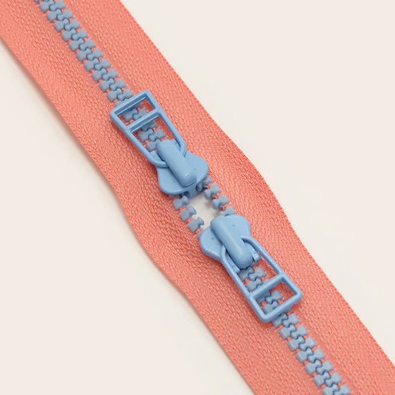 May include: A close-up of a pink zipper with two blue plastic zipper pulls. The zipper is open and the pulls are visible.