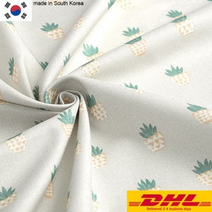 Op de afbeelding: Witte stof met een herhalend patroon van groene en gele ananas. De stof is gemaakt in Zuid-Korea. De tekst "DHL Geleverd in 2-4 werkdagen" staat onderaan de afbeelding.