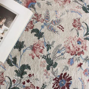 May include: A beige floral fabric with a detailed pattern of pink, red, and blue flowers and green leaves. The design includes a variety of floral elements and winding stems. A book with a white cover is partially visible in the upper left corner.