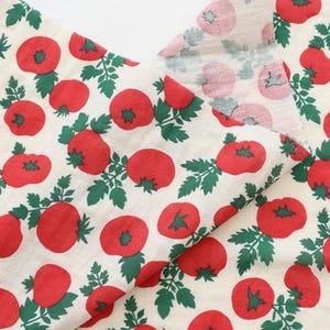 May include: A piece of off-white fabric featuring a repeating pattern of bright red tomatoes with green stems and leaves. The design is printed across the textile, showing the front side and a folded corner revealing the reverse side of the fabric.