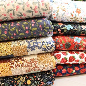 Könnte beinhalten: Ein Stapel gerollter, gesteppter Stoffe mit verschiedenen Blumen- und Patchworkmustern. Die Stoffe zeigen Designs mit rosa Blumen, Erdbeeren und anderen botanischen Motiven in Blau, Rot, Gelb und Weiß. Die Quilts sind ordentlich gestapelt.