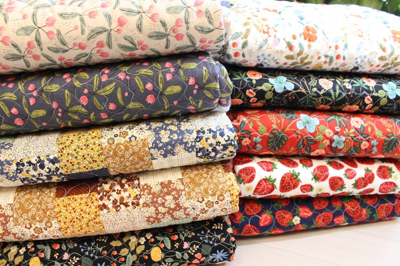 Pu&ograve; includere: Una pila di tessuti trapuntati piegati con vari motivi, tra cui stampe floreali rosa su crema, fiori blu scuro, quadrati patchwork in giallo e marrone e motivi a fragole rosse su sfondi bianchi e blu scuro.