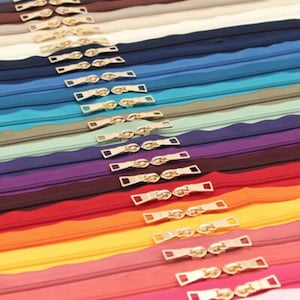 Può includere: Una collezione di cerniere colorate in varie tonalità, tra cui bianco, blu navy, blu, verde acqua, grigio, viola, rosso, arancione e rosa. Ogni cerniera ha un tirante dorato. Le cerniere sono disposte in file parallele.