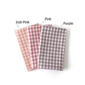 May include: Three folded fabric swatches in Indi-Pink, Pink, and Purple. Each swatch features a classic gingham check pattern. The fabric is arranged on a white background, with the color names labeled above each swatch.