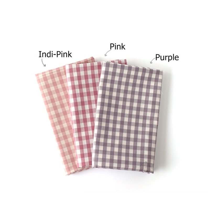 May include: Three folded fabric swatches in Indi-Pink, Pink, and Purple. Each swatch features a classic gingham check pattern. The fabric is arranged on a white background, with the color names labeled above each swatch.