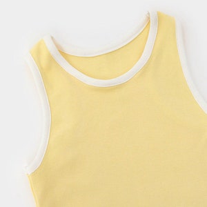 Pu&ograve; includere: Una canotta senza maniche di colore giallo pastello tenue con un bordo a coste bianco a contrasto lungo lo scollo rotondo e gli scalfi manica. Il capo &egrave; realizzato in un tessuto liscio a tinta unita con una struttura semplice.