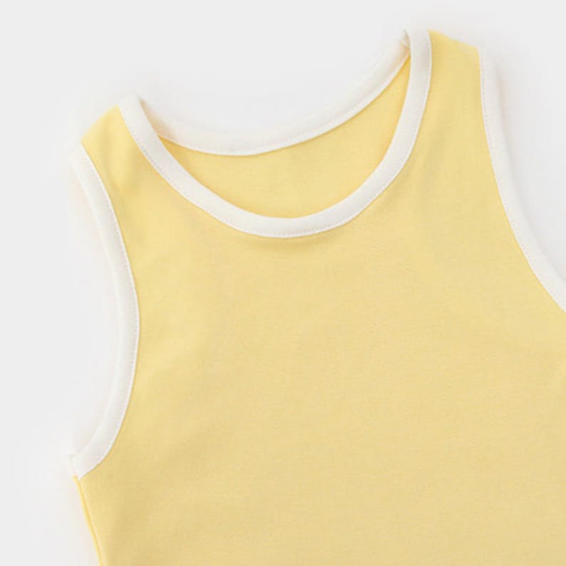 Pu&ograve; includere: Una canotta senza maniche di colore giallo pastello tenue con un bordo a coste bianco a contrasto lungo lo scollo rotondo e gli scalfi manica. Il capo &egrave; realizzato in un tessuto liscio a tinta unita con una struttura semplice.