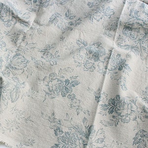 K&ouml;nnte beinhalten: Ein St&uuml;ck cremefarbener Stoff mit einem sich wiederholenden Blumenmuster in einem ged&auml;mpften Blauton. Das Design besteht aus detaillierten Illustrationen von bl&uuml;henden Blumen, Bl&auml;ttern und St&auml;ngeln, die &uuml;ber die strukturierte Oberfl&auml;che verteilt sind.