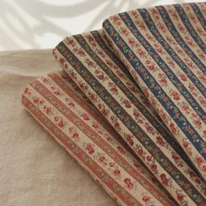 May include: Close-up of three fabric rolls with vertical stripes. The stripes alternate between floral patterns and solid colors, including red, blue, and brown. The floral stripes feature small red flowers and green leaves.