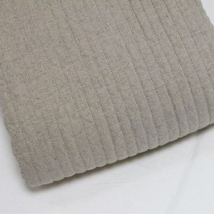 May include: A close-up of a beige ribbed fabric with a subtle sheen. The fabric is folded and has a soft, textured appearance.