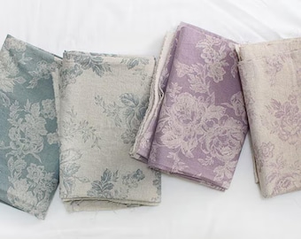 May include: Four folded fabric squares in shades of blue, grey, lavender, and beige, each with a floral pattern. The fabrics are arranged on a white surface, showcasing the intricate design and colour variations.