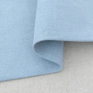 May include: Close-up of a light blue fabric with a textured weave. The fabric is folded over, revealing a curved edge. The background is a neutral, off-white fabric. The image highlights the texture and color of the textile.
