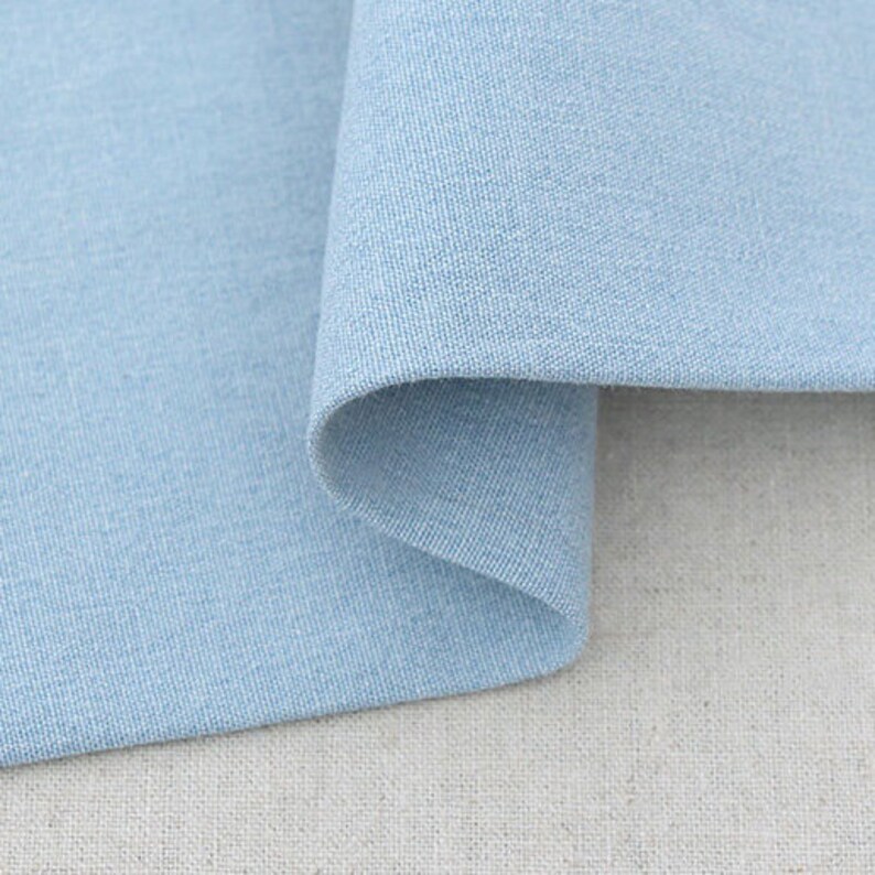 May include: Close-up of a light blue fabric with a textured weave. The fabric is folded over, revealing a curved edge. The background is a neutral, off-white fabric. The image highlights the texture and color of the textile.