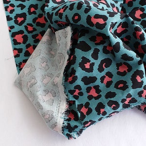 May include: Teal fabric with a pink and black leopard print pattern. The fabric is folded and the edges are frayed.