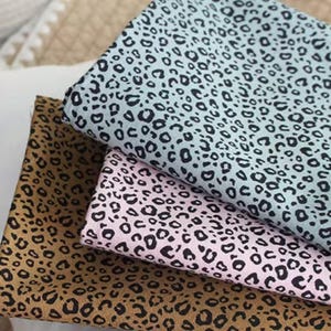 May include: Three fabric swatches with a leopard print pattern. The colours are tan, light pink, and light blue, each with black spots. The fabric is folded, showing the pattern and texture of the material. The swatches are stacked on top of each other.