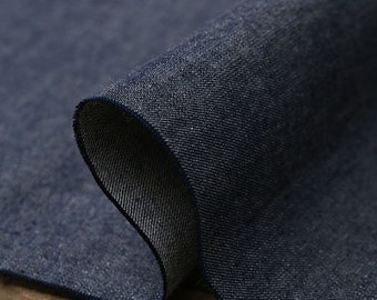 May include: Close-up of a rolled section of dark blue denim fabric. The fabric has a textured surface with visible weave detail. The edges of the fabric are visible, showing the layers and the contrasting lighter underside.