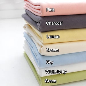 May include: A pile of folded fabric in shades of pink, charcoal, lemon, cream, sky blue, white-ivory, and green. Each fabric is labelled with its colour name. The fabrics are neatly stacked, displaying the range of available colours.