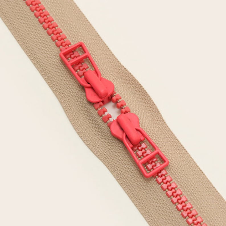 May include: A beige zipper with two pink plastic sliders. The zipper is closed and the sliders are in the middle of the zipper.