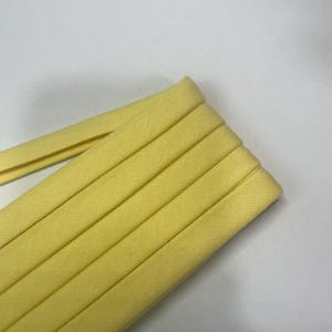 Pu&ograve; includere: Un rotolo di nastro sbieco giallo pallido caratterizzato da una trama di tessuto intrecciato. Il nastro &egrave; piegato ordinatamente in pi&ugrave; strati, mostrando una larghezza costante e una finitura liscia e opaca lungo tutta la lunghezza del materiale.