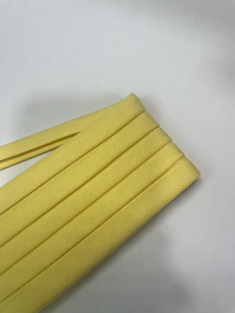 Pu&ograve; includere: Un rotolo di nastro sbieco giallo pallido caratterizzato da una trama di tessuto intrecciato. Il nastro &egrave; piegato ordinatamente in pi&ugrave; strati, mostrando una larghezza costante e una finitura liscia e opaca lungo tutta la lunghezza del materiale.