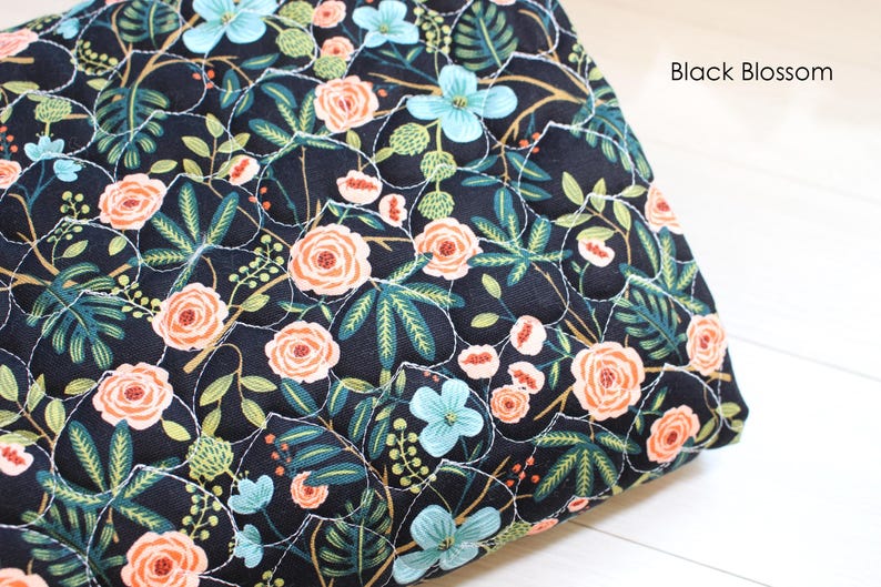 Pu&ograve; includere: Un tessuto trapuntato con sfondo nero e un motivo floreale di rose color pesca, fiori azzurri e fogliame verde scuro. Il tessuto presenta un motivo a griglia cucito in bianco e il testo Black Blossom nell'angolo.