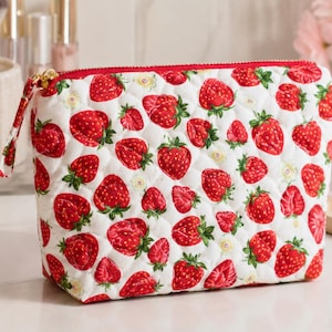 Pu&ograve; includere: Una pochette in tessuto trapuntato con un motivo ripetuto di fragole rosse e piccoli fiori bianchi su sfondo bianco. La pochette presenta una chiusura a zip rossa sulla parte superiore e una linguetta in tessuto abbinata, che conferisce una forma strutturata.