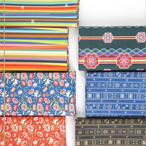 Puede incluir: Una colección de seis muestras de tela plegadas con diversos patrones. Dos muestras muestran rayas horizontales en colores vibrantes. Dos presentan motivos florales y de grullas en azul y rojo. Dos muestran patrones geométricos con símbolos tradicionales.