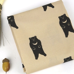 Puede incluir: Tela beige con un patrón repetido de osos negros. Los osos están estilizados y tienen una forma de media luna en el pecho.