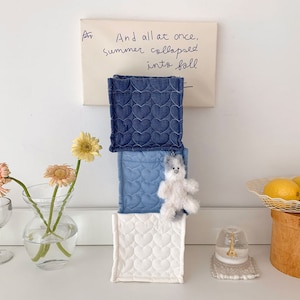 May include: Three square fabric containers stacked vertically, each with a heart-shaped quilted pattern. The top container is dark blue, the middle is light blue, and the bottom is white. A small, fluffy bear keychain hangs from the middle container. A handwritten note is above the containers.