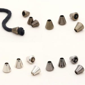 May include: Various metal cone-shaped cord ends in silver, bronze, and gunmetal finishes. One is attached to a dark blue rope. These findings are used for jewellery making and crafts. Measurements are in centimetres.