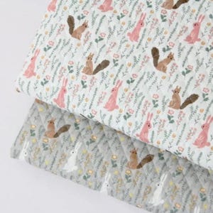 May include: A quilted fabric featuring a repeating pattern of brown squirrels and pink rabbits interspersed with green foliage and small flowers. The top layer is white, while the bottom layer is a light grey, both with a diamond-stitched texture.