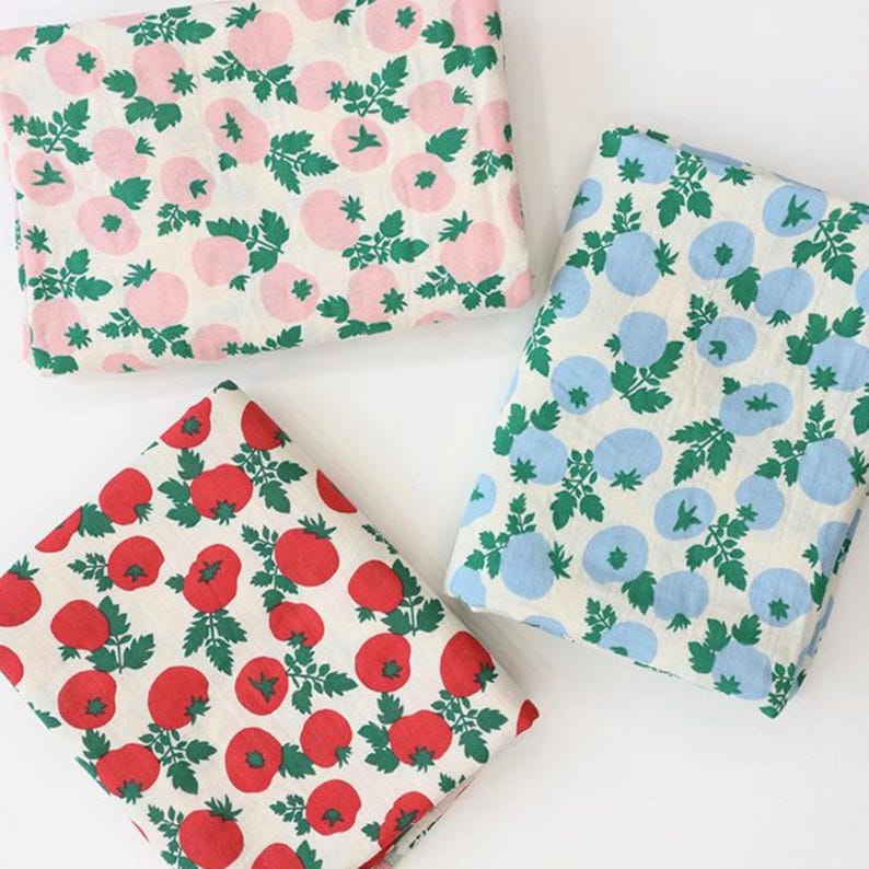 May include: Three folded pieces of fabric featuring a repeating pattern of round fruits and green leaves. One piece has light pink fruits on a white background, one has bright red fruits on a white background, and one has light blue fruits on a white background.