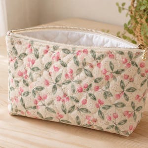 Pu&ograve; includere: Una pochette in tessuto trapuntato con base color crema e un motivo ripetuto di piccoli fiori rosa e foglie verdi. La borsa rettangolare presenta una chiusura a zip in metallo dorato e una fodera interna bianca trapuntata visibile all'apertura.