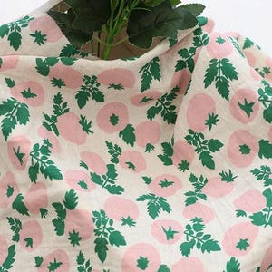 May include: A piece of off-white fabric featuring a repeating pattern of light pink circular shapes and dark green leafy stems. The textile has a soft, draped appearance with a visible weave, showcasing a botanical-inspired print throughout.