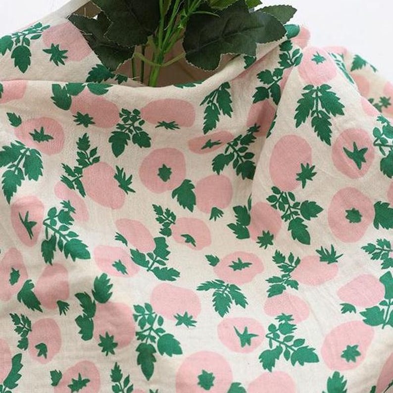 May include: A piece of off-white fabric featuring a repeating pattern of light pink circular shapes and dark green leafy stems. The textile has a soft, draped appearance with a visible weave, showcasing a botanical-inspired print throughout.