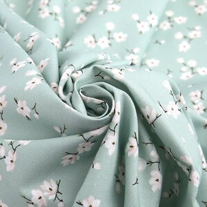 May include: A light mint green fabric featuring a repeating pattern of small white flowers with dark brown stems and centers. The textile is gathered in the center, creating soft folds and shadows that highlight the texture of the woven material.