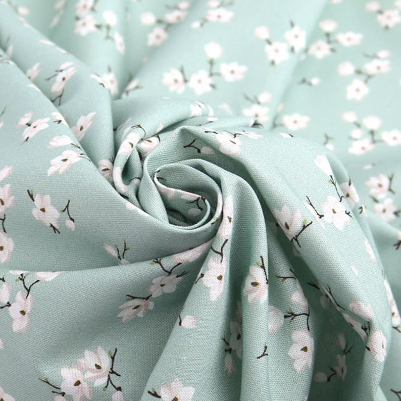 May include: A light mint green fabric featuring a repeating pattern of small white flowers with dark brown stems and centers. The textile is gathered in the center, creating soft folds and shadows that highlight the texture of the woven material.