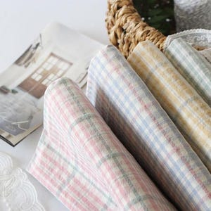 Peut inclure: Un ensemble de quatre serviettes en tissu pliées présentant un motif à carreaux vichy doux dans des tons pastel de rose, bleu, jaune et vert. Les textiles sont disposés dans un panier tressé à côté d'un petit livret imprimé et de dentelle blanche.