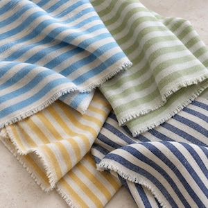 May include: A collection of four folded fabric pieces featuring vertical stripes in light blue, sage green, mustard yellow, and navy blue. Each piece of textile displays a natural, raw-edged fringe along the borders, showcasing a woven texture.
