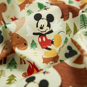 May include: A cream-colored fabric featuring a repeating pattern of Mickey Mouse standing on a tree stump, brown moose, green pine trees, red campfires, and small colorful triangular bunting flags printed across the surface of the material.