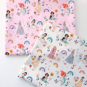 Puede incluir: Dos piezas de tela con fondo rosa y blanco que presentan un patrón repetido de princesas Disney, arcoíris y flores. Las princesas incluyen Cenicienta, Ariel, Jasmine, Rapunzel y Blancanieves.