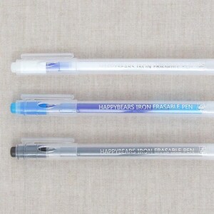 May include: Three clear plastic pens with the text "HAPPYBEARS IRON ERASABLE PEN". The pens have white, blue, and grey accents. The pens are lying on a light beige fabric.