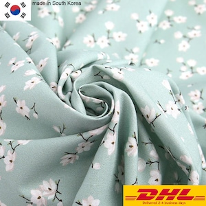 May include: A light mint green fabric featuring a repeating pattern of small white cherry blossom flowers with dark brown stems. The textile is gathered in the center, creating soft folds and shadows that highlight the texture of the material.