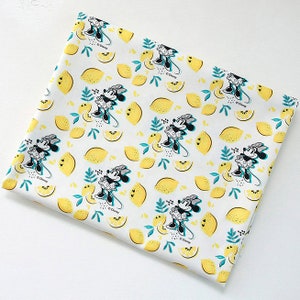 Puede incluir: Tela blanca con un patr&oacute;n repetido de Minnie Mouse y limones. Minnie Mouse lleva un vestido blanco y negro con un lazo rojo. Los limones son amarillos con hojas verdes. La tela tiene un fondo blanco con una textura sutil.
